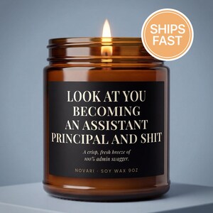 May include: A brown glass jar candle with a lit wick and a black label featuring the white text: LOOK AT YOU BECOMING AN ASSISTANT PRINCIPAL AND SHIT. Below this, it reads: A crisp, fresh breeze of 100% admin swagger. NOVARI SOY WAX 9OZ.