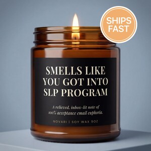 May include: A lit candle in a brown glass jar with a black label. The label reads "SMELLS LIKE YOU GOT INTO SLP PROGRAM." Below the text is "A relieved, inbox-lit note of 100% acceptance email euphoria." The jar is 9oz.