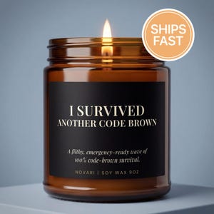 May include: A lit brown glass candle jar with a black label that reads "I SURVIVED ANOTHER CODE BROWN". The candle is labeled "NOVARI | SOY WAX 9OZ". A round orange sticker with the words "SHIPS FAST" is on the top right.