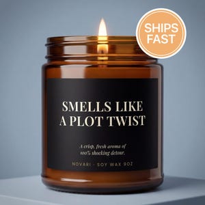 May include: A lit candle in an amber glass jar with a black label featuring the text "SMELLS LIKE A PLOT TWIST" in white, along with "A crisp, fresh aroma of 100% shocking detour," "NOVARI," and "SOY WAX 9OZ" printed on the front of the container.