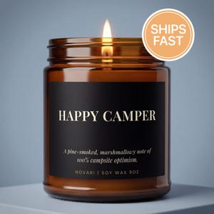 May include: A lit candle in an amber glass jar with a black label. The label reads HAPPY CAMPER, A pine-smoked, marshmallowy note of 100% campsite optimism, NOVARI | SOY WAX 9OZ. The candle features a single flame and is contained in a 9oz glass vessel.