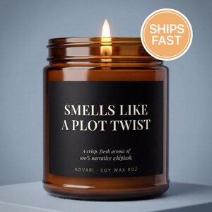 May include: A lit candle in an amber glass jar with a black label featuring the text "SMELLS LIKE A PLOT TWIST" in white, followed by "A crisp, fresh aroma of 100% narrative whiplash." and "NOVARI · SOY WAX 9OZ" printed at the bottom.