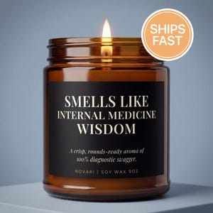 May include: A lit candle in an amber glass jar with a black label. The label reads: SMELLS LIKE INTERNAL MEDICINE WISDOM, A crisp, rounds-ready aroma of 100% diagnostic swagger, NOVARI | SOY WAX 9OZ. The candle features a single burning wick.