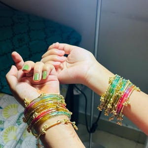 May include: Two wrists displaying stacks of vibrant bangles. The bangles are a mix of transparent and solid colours, including red, yellow, green, and pink. Gold-coloured accents and embellishments add detail to the jewellery.