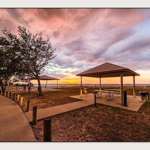 May include: A park scene featuring two metal-roofed pavilions with picnic tables, situated on concrete pads near a shoreline. A paved walkway curves through the grassy area, lined with wooden posts, under a sky filled with pink and orange sunset clouds.