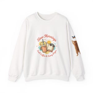 Op de afbeelding: Witte sweatshirt met een grafisch ontwerp met de tekst "Slow Mornings", een ijskoffie, een zonnebril en een boek. Een cartoonkat en een vlinder staan op de rechter mouw. Het ontwerp is in tinten oranje, geel en bruin.