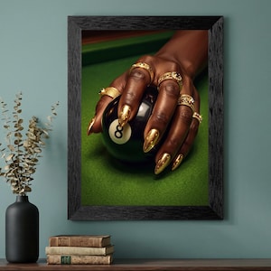 May include: Framed print featuring a close-up of a hand with gold-colored nails and rings holding a black eight-ball on a green felt surface. The image is set in a black frame.