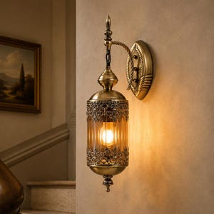 Puede incluir: Un aplique de pared con acabado en latón antiguo, una pantalla cilíndrica de cristal estriado y detalles metálicos de filigrana. La lámpara cuelga de un brazo curvo sujeto a una placa de pared ovalada, emitiendo una luz cálida.