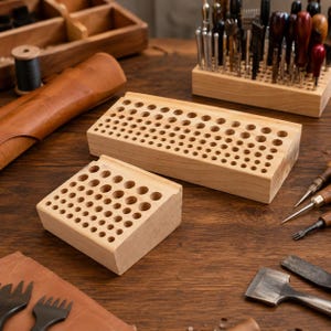 May include: Two light-colored wooden tool organizers with multiple circular holes of varying diameters arranged in rows. These rectangular blocks are designed to hold various leatherworking tools and punches in an upright position.
