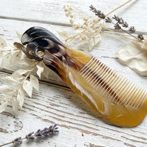May include: A decorative comb with a carved feather design. The comb is made of a light amber-coloured material with dark brown and black accents. The comb is approximately 15 cm long. The background includes white flowers and lavender sprigs.