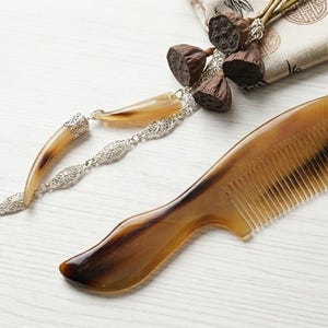 Puede incluir: Un peine de cuerno marrón con mango curvo y dientes finos, junto a una cadena decorativa plateada con colgantes en forma de cuerno y una pequeña bolsa con vainas de semillas secas. El peine está hecho de un material natural.