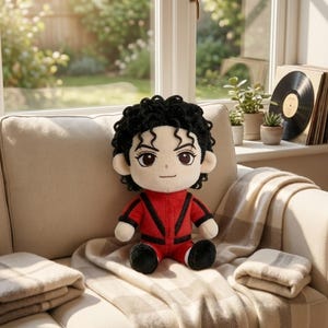 May include: A plush doll featuring a person with curly black hair, large brown eyes, and a light skin tone. The doll wears a red jacket with black V-shaped accents and matching red pants with black cuffs, sitting in a seated position.