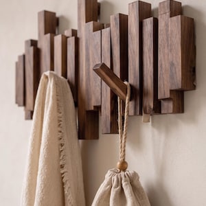 May include: A wall-mounted coat rack constructed from vertical dark brown wooden slats of varying heights. The rack features a single protruding wooden hook holding a beige drawstring pouch, while a beige fringed towel hangs from another section.