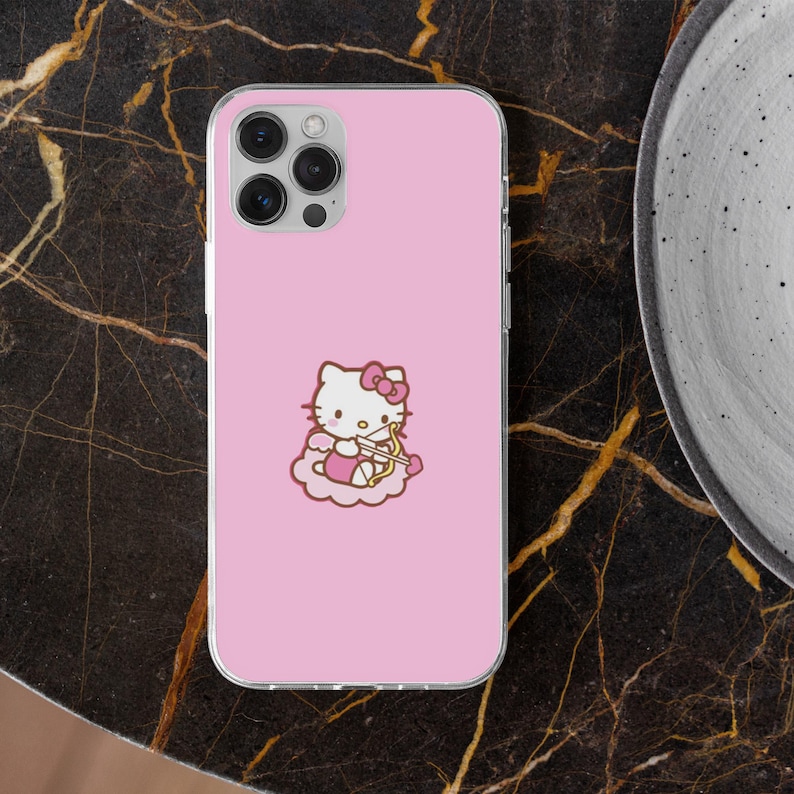 May include: A pink phone case featuring a cartoon image of Hello Kitty as Cupid, complete with a bow and arrow, sitting on a cloud. The case has a clear border and is set against a dark, marble-like surface.