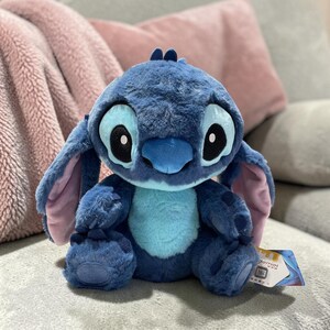 Puede incluir: Un peluche del personaje Stitch de la pel&iacute;cula de Disney Lilo & Stitch. El juguete es principalmente azul con grandes ojos negros, una barriga azul claro y orejas internas rosas. Est&aacute; sentado sobre una superficie gris.
