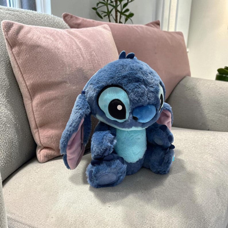 Puede incluir: Un peluche azul de Stitch con grandes ojos negros, una barriga azul claro y orejas grandes. El peluche est&aacute; sentado en un sof&aacute; gris con cojines rosas en el fondo. El mu&ntilde;eco es un personaje de la pel&iacute;cula animada Lilo & Stitch.