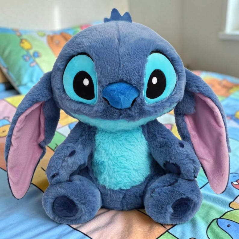 Puede incluir: Un peluche azul de un personaje alien&iacute;gena con ojos y orejas grandes. El juguete tiene una panza azul claro y orejas internas rosas. Est&aacute; sentado sobre una colcha estampada.