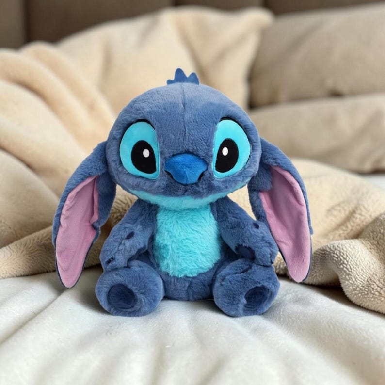 Puede incluir: Un peluche azul del personaje Stitch de la pel&iacute;cula animada Lilo & Stitch. El juguete tiene grandes ojos ovalados, una nariz azul y orejas grandes con el interior rosa. Est&aacute; sentado sobre una manta blanca.