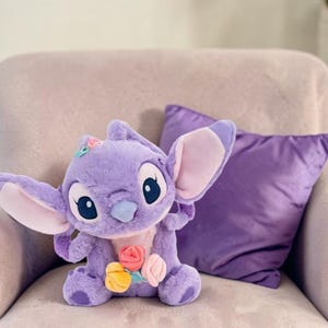 Puede incluir: Un personaje de peluche de color lavanda con orejas grandes, ojos grandes y una nariz azul. El peluche tiene una peque&ntilde;a flor en la cabeza y sostiene un ramo de flores coloridas. Una almohada morada descansa detr&aacute;s sobre una silla de color claro.