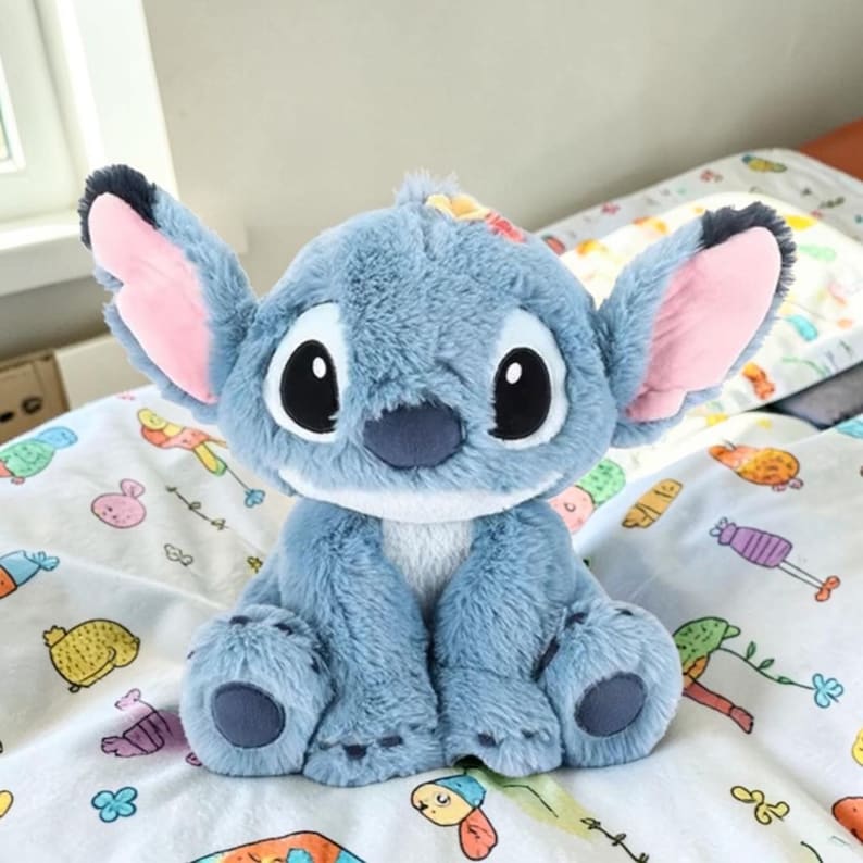Puede incluir: Un peluche azul del personaje Stitch, con grandes ojos negros, orejas rosas y vientre blanco. La criatura peluda est&aacute; sentada sobre una s&aacute;bana blanca con un estampado colorido. Un juguete popular para ni&ntilde;os.