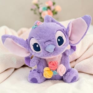 Puede incluir: Un peluche morado de un personaje de dibujos animados con grandes orejas y ojos azules, sosteniendo un ramo de flores. El juguete tiene una peque&ntilde;a flor en la cabeza. El fondo es borroso.