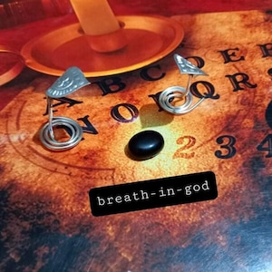 May include: Two silver-toned metal planchette-shaped charms with spiral bases and a single smooth black oval stone. The charms feature embossed sun and moon motifs. A black rectangular label with white text reads breath-in-god.