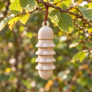 Op de afbeelding: Een lichtbeige, gelaagd, cilindervormig hangend ornament met een gladde, matte afwerking. Het object heeft vier afzonderlijke, afgeronde, bloembladachtige lagen die verticaal zijn gestapeld en opgehangen aan een bruin leren koord.