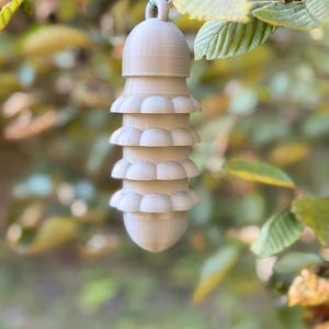 May include: A light brown, vertically oriented, cylindrical ornament featuring a smooth, rounded top with a small hanging loop and three distinct, stacked, scalloped-edge tiers, ending in a smooth, rounded, tapered base point.