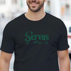 May include: A black short-sleeved t-shirt featuring the word Servus printed in a dark green font across the chest. Below the text is a small graphic of mountain peaks flanked by two snowflake-like symbols, all rendered in the same dark green color.