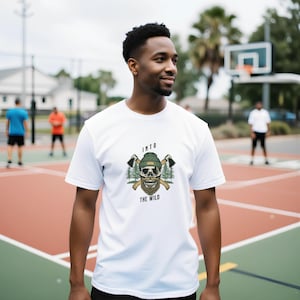 Könnte beinhalten: Weißes T-Shirt mit einer Grafik eines Schädels, der eine grüne Mütze trägt und gekreuzte Äxte hält, mit dem Text "INTO THE WILD". Das Design beinhaltet Bäume und einen Bart. Das T-Shirt wird von einer Person auf einem Basketballplatz getragen.
