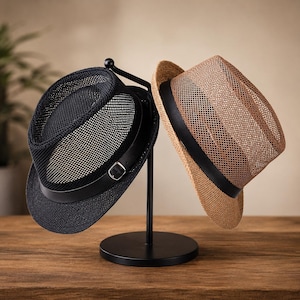 Op de afbeelding: Een zwarte mesh fedora en een beige mesh fedora op een zwarte metalen standaard. De zwarte hoed heeft een zwarte band met een zilverkleurige metalen gesp. Beide hoeden hebben een gestructureerde rand en een geweven mesh kroonontwerp.