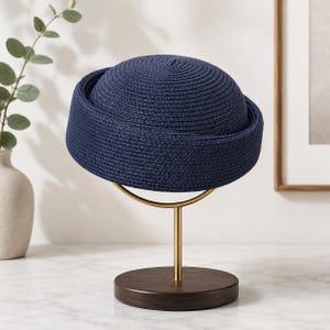 Peut inclure: Un chapeau en paille bleu marine doté d'une calotte arrondie et d'un bord structuré retroussé. Le chapeau est présenté sur un support en métal doré composé d'un anneau de maintien circulaire et d'une base ronde en bois foncé.