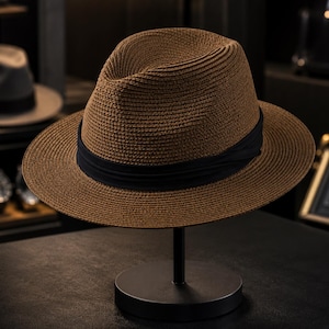 Pode incluir: Um chapéu fedora castanho com uma aparência texturizada e entrelaçada, apresentando uma coroa vincada e uma aba larga. Uma faixa de tecido preto sólido envolve a base da coroa, proporcionando um contraste distinto com o material castanho.