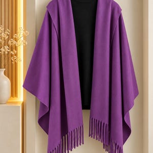 May include: A vibrant purple hooded poncho featuring an open front design and long fringe detailing along the bottom hem, layered over a solid black mock neck sweater, all displayed on a wooden hanger against a neutral wall.