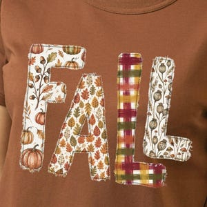 May include: A brown short-sleeved t-shirt featuring the word FALL in large block letters. The F is decorated with pumpkins and acorns, the A with autumn leaves, the L with a red and green plaid pattern, and the second L with dried flowers.