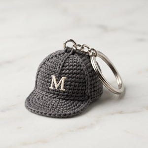 Könnte beinhalten: Ein grauer gehäkelter Baseballkappen-Schlüsselanhänger mit einem silbernen Schlüsselring. Die Kappe hat einen weißen, gestickten Buchstaben "M" auf der Vorderseite. Der Schlüsselanhänger ist eine Miniaturversion einer Baseballkappe.