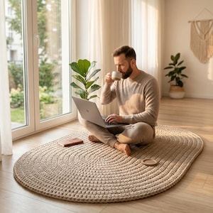 May include: An oval-shaped, beige chunky knit rug with a thick, braided texture. The rug is placed on a light-colored wooden floor, providing a soft surface for sitting while using a silver laptop and holding a small, plain white ceramic mug.