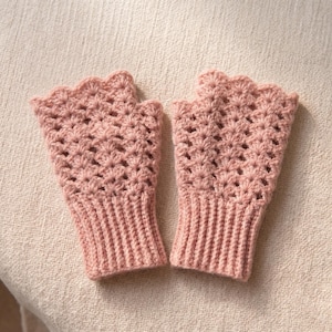 Könnte beinhalten: Ein Paar rosa fingerlose Handschuhe. Die Handschuhe bestehen aus gehäkeltem Material mit einem gewellten Rand. Die Handgelenkbündchen sind gerippt. Die Handschuhe werden auf einer neutralfarbenen Oberfläche präsentiert.