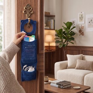 May include: A navy blue crochet door organizer with three pockets, hanging from a decorative brass hook. The top pocket holds a smartphone displaying an app, while the lower pockets contain white charging cables and a power adapter. The organizer is designed for home organization.