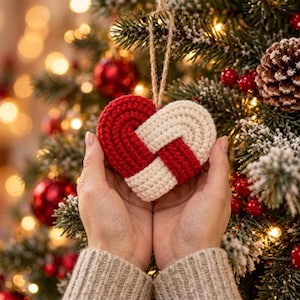 May include: A heart-shaped ornament crafted from crocheted yarn in alternating red and cream sections. The heart features an interlocking woven design and is suspended by a thin, light brown jute twine loop for hanging on a tree branch.