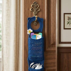 May include: A blue crochet phone organizer with a flower accent, hanging on a wooden door. The organizer has a top ring for hanging, a pocket for a phone, and a lower pocket for charging cables and a power adapter. The phone displays the weather.