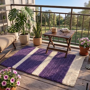 Può includere: Un tappeto all'uncinetto a righe viola e bianche su un terrazzo in legno. Il tappeto è rettangolare e presenta due ampie strisce bianche. Il terrazzo comprende anche un tavolino in legno, piante in vaso e una sedia.
