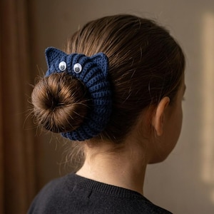 Könnte beinhalten: Ein dunkelblaues gehäkeltes Haargummi mit zwei spitzen Katzenohren und zwei weißen Wackelaugen mit schwarzen Pupillen. Das Accessoire ist um einen braunen Haarknoten gewickelt und erzeugt durch die Textur des Garns eine katzenartige Optik.