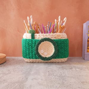 Puede incluir: Un organizador hecho a mano con forma de cámara de ganchillo en verde y crema, lleno de ganchos de ganchillo. La cámara tiene un diseño de lente circular y un pequeño botón verde. Una planta suculenta y una pantalla iluminada en miniatura están en el fondo.