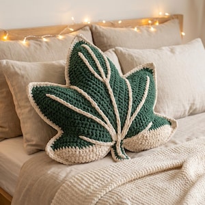 May include: A handmade, green leaf-shaped pillow with cream-colored accents rests on a bed. The pillow is made of a textured material and has a detailed leaf vein design. The bed is made up with neutral-colored linens.