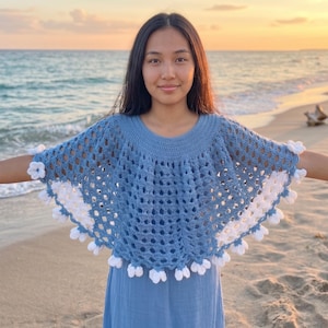 May include: A light blue crochet poncho with white flower accents. The open weave design is visible, and the poncho is worn over a blue dress. The background shows a beach and ocean at sunset.