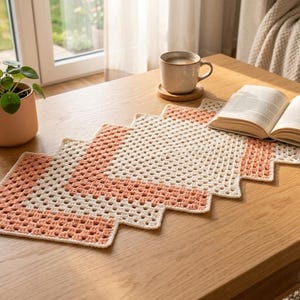 May include: A handmade crochet table runner in a zig-zag pattern. The runner features alternating stripes of cream and peach colors. A cup of coffee, a book, and a potted plant are on the wooden table.