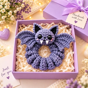 Op de afbeelding: Een paarse gehaakte vleermuis-scrunchie met zwarte binnenoren, grote zwarte veiligheidsogen, roze wangen en twee witte hoektanden. De vleermuis ligt in het midden van een vierkante paarse geschenkdoos gevuld met versnipperd papier.