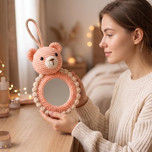 May include: A handmade, peach-colored crochet mirror with a bear-shaped top and a loop handle. The mirror has a round shape with a decorative border. The bear has a cream-colored snout and a small, dark-colored nose and eyes. The mirror is approximately 5 inches in diameter.