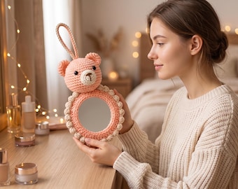 May include: A handmade, peach-colored crochet mirror with a bear-shaped top and a loop handle. The mirror has a round shape with a decorative border. The bear has a cream-colored snout and a small, dark-colored nose and eyes. The mirror is approximately 5 inches in diameter.
